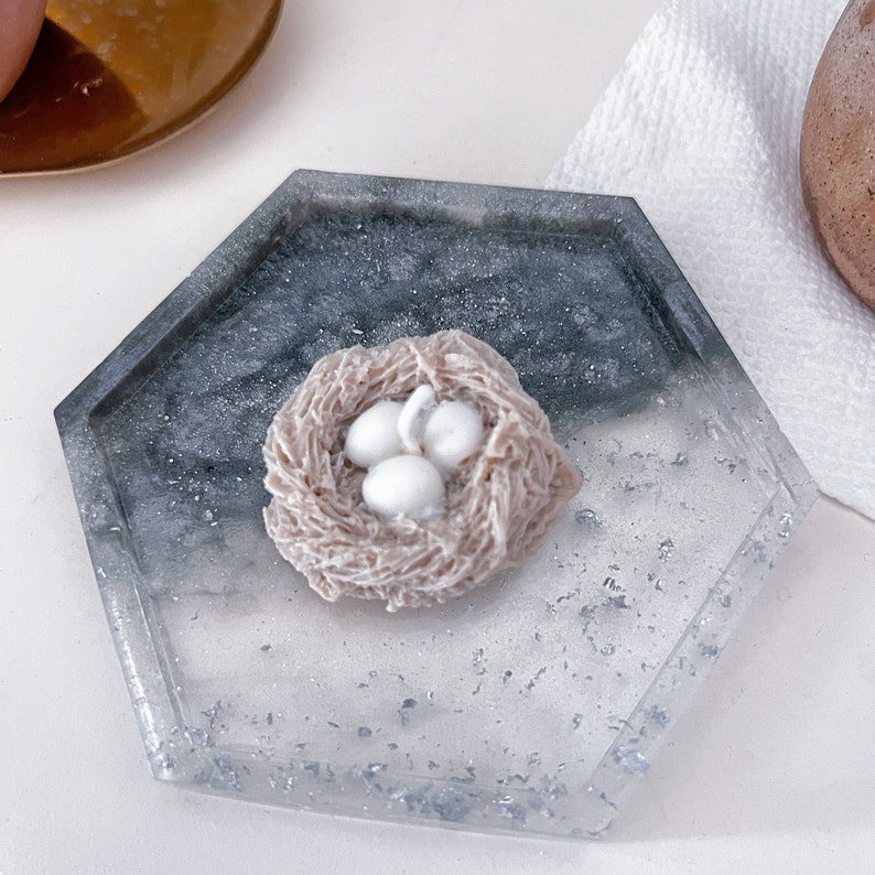 May include: A white ceramic bird's nest candle with three white eggs inside. The nest is sitting on a gray and white hexagonal tray with a glittery finish.