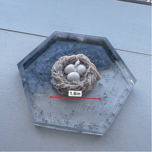 May include: A white ceramic bird's nest with three white eggs inside, sitting on a gray and white speckled tray. The nest is 1.8 inches wide.