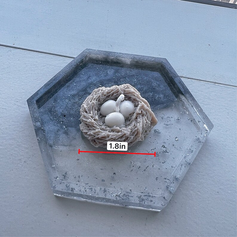 May include: A white ceramic bird's nest with three white eggs inside, sitting on a gray and white speckled tray. The nest is 1.8 inches wide.