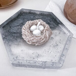 May include: A white candle shaped like a bird's nest with three white eggs inside. The candle is sitting on a gray and white tray.