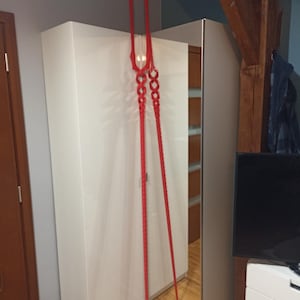 May include: A red spear-like decorative object hangs in front of a white wardrobe with a mirrored door. The object has a central head with a series of connected rings and two long, pointed shafts. The background includes a wooden beam and a television.