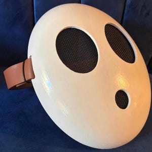 Könnte beinhalten: Eine weiße, ovale Maske mit drei schwarzen, mit Mesh bedeckten Löchern. Auf der Seite ist ein brauner Lederriemen befestigt. Diese Maske ist wahrscheinlich ein Cosplay- oder Kostümzubehör.