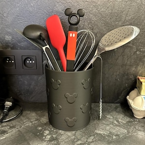 May include: A black utensil holder with embossed Mickey Mouse silhouettes, filled with various kitchen tools. Includes a red spatula, whisk, and slotted spoon. The holder sits on a dark countertop.