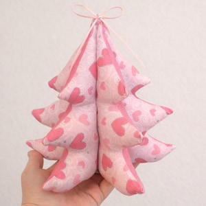 Könnte beinhalten: Ein rosa Weihnachtsbaum aus Stoff mit einem Herzmuster. Der Baum besteht aus weichem, gepolstertem Material und hat ein hellrosa Band, das oben gebunden ist. Der Baum wird in der Hand einer Person gehalten.