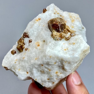 May include: A raw, natural mineral specimen featuring a large, brown crystal cluster on a white matrix. Smaller, scattered crystals of the same color are visible. The stone has a rough, textured surface with orange and brown spots.
