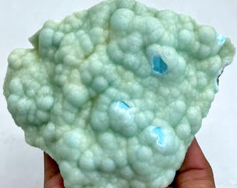 Op de afbeelding: Een lichtgroen mineraal specimen met een bubbelige, getextureerde oppervlakte. Het mineraal heeft kleine zakken met helderblauwe kleur. De algemene vorm is onregelmatig en organisch, met een enigszins ruwe textuur.