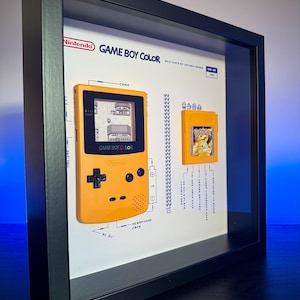 Può includere: Una teca incorniciata di un Game Boy Color giallo con una cartuccia Pokemon. Il display include uno schema delle caratteristiche del Game Boy Color, tra cui il jack per le cuffie, il controllo del volume e il pulsante di accensione.