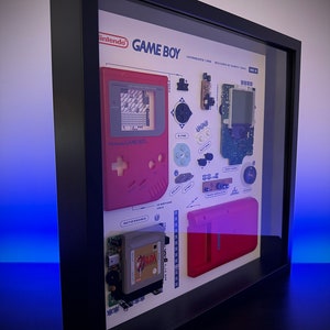 Può includere: Una vetrina incorniciata di un Game Boy smontato, che mostra i singoli componenti del dispositivo di gioco portatile. La vetrina include il Game Boy originale, una cartuccia e vari pulsanti e pezzi. Il testo "Nintendo GAME BOY INTRODUCED 1989 DESIGNED BY GUMPEI YOKOI" è visibile nella parte superiore della vetrina.