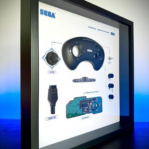 May include: Framed print featuring a Sega Mega Drive controller and internal components. The print includes a black controller, D-pad, connector, and motherboard, with the Sega logo in blue. The print is encased in a black frame.