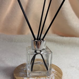May include: A clear square glass reed diffuser bottle with a silver collar and four black reeds. The bottle sits on a round light wood coaster featuring a white floral pattern printed on the surface. The design is simple and functional.