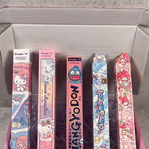 Può includere: Una collezione di cinque astucci colorati con personaggi Sanrio, tra cui Hello Kitty, Hangyodon e Cinnamoroll, all'interno di una scatola a scacchi rosa e bianca. Gli astucci contengono testo in inglese e cinese.