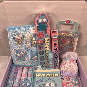 Pode incluir: Uma caixa de presente rosa e branca cheia de vários artigos de papelaria com um personagem de desenho animado azul chamado Hangyodon. Os artigos incluem adesivos, uma régua, um lápis, um apontador, um bloco de notas e um pequeno saco de borrachas. A caixa é decorada com o logotipo da Sanrio.