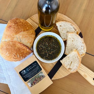 May include: A wooden cutting board with a black stripe, a bowl of green olive oil dip, a bottle of olive oil, and four slices of white bread. A small brown paper bag with the text "Sunshine in Every Bread Dip Seasoning" is on a white cloth.