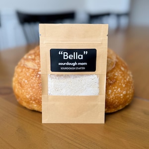May include: A small brown paper pouch with a white label that says "Bella" sourdough mom sourdough starter. The pouch is sitting on a wooden table next to a loaf of sourdough bread.