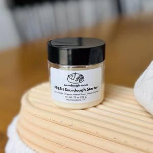 May include: A small, clear jar with a black lid, labeled "FRESH Sourdough Starter." The label includes the ingredients: organic wheat flour and filtered water. The jar is .75 oz (20 g) and sits on a wooden surface.