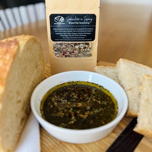 May include: A rustic bread dip set featuring a white bowl of olive oil and herbs, a loaf of bread, and a packet of "Somewhere in Tuscany" bread dip seasoning. A bottle of olive oil is in the background. The seasoning packet includes the ingredients.