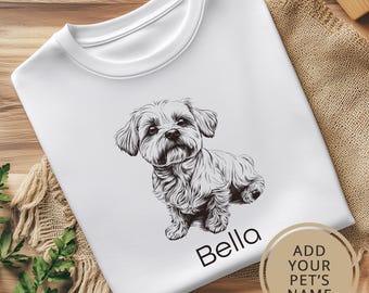 May include: White t-shirt with a black and white illustration of a small dog sitting. The dog's name, "Bella", is printed below the illustration. The text "ADD YOUR PET'S NAME" is printed in a circle on the bottom right corner of the shirt.