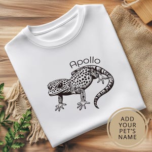 K&ouml;nnte beinhalten: Wei&szlig;es T-Shirt mit einer schwarz-wei&szlig;en Illustration eines Gecko-Leguan. Der Text "Apollo" ist &uuml;ber dem Leguan gedruckt.