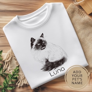 May include: White t-shirt with a black and white illustration of a cat. The cat is sitting with its tail curled around its body. The text "Luna" is printed below the cat. The text "Add your pet's name" is printed in a circle on the bottom right corner of the shirt.