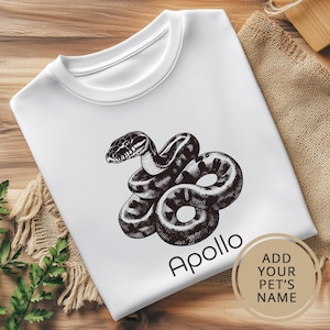 May include: White t-shirt featuring a black and white illustration of a snake. The text "Apollo" is printed below the snake. The text "ADD YOUR PET'S NAME" is printed in a circle on the bottom right corner of the shirt.