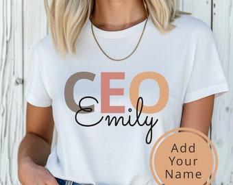 May include: White t-shirt with the text "CEO Emily" in brown, pink, and orange lettering. The text "Add Your Name" is written in a circle on the bottom right corner of the shirt.