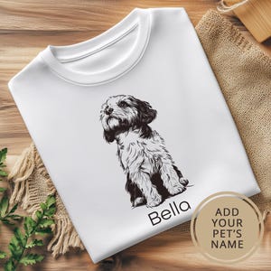 May include: White t-shirt with a black and white illustration of a dog sitting. The text "Bella" is printed below the dog. The text "ADD YOUR PET'S NAME" is printed in a circle on the bottom right corner of the shirt.