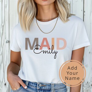 以下が含まれることがあります： 「MAID Emily」という文字が茶色、ピンク、オレンジの文字で書かれた白いTシャツ。画像の右下隅には円の中に「Add Your Name」という文字が書かれている。