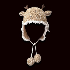 May include: A crocheted reindeer hat in tan and white, featuring a deer head design with antlers and ear flaps. The hat has a white lace trim and two pom-poms hanging from the ear flaps. The hat is designed to be worn on the head.