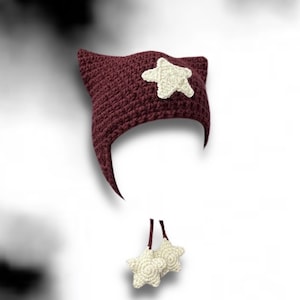 May include: A maroon crocheted hat with cat ears and a white star on the front. The hat has two white star-shaped charms hanging from the bottom.