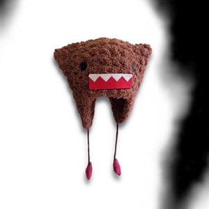 Pode incluir: Um gorro de crochê castanho com textura, apresentando uma boca retangular com dentes brancos em ziguezague sobre um fundo vermelho. O gorro tem dois olhos pretos circulares e dois cordões finos castanhos pendentes com pequenos detalhes vermelhos.