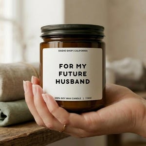 May include: A brown glass candle jar with a black lid, held in a hand. The label reads "FOR MY FUTURE HUSBAND" in bold black letters. The candle is 7.5 oz and made of 100% soy wax.