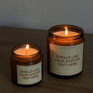 Peut inclure: Deux bougies en pot en verre ambr&eacute; avec des m&egrave;ches allum&eacute;es. Chaque bougie pr&eacute;sente une &eacute;tiquette cr&egrave;me avec le texte "SMELLS LIKE YOUR CUSTOM TEXT HERE", "DASHIO SHOP", "MADE IN CALIFORNIA", "COFFEE SHOP", "100% SOY WAX CANDLE" et "7.5 OZ".