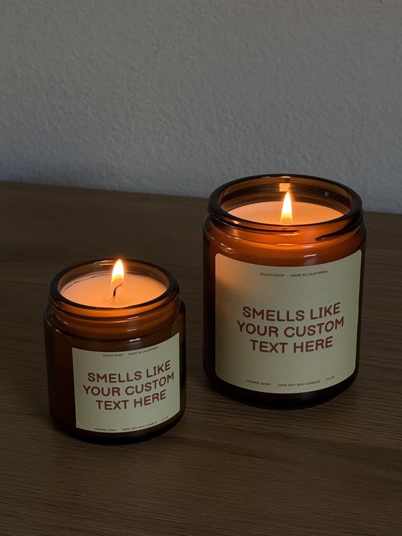 Peut inclure: Deux bougies en pot en verre ambr&eacute; avec des m&egrave;ches allum&eacute;es. Chaque bougie pr&eacute;sente une &eacute;tiquette cr&egrave;me avec le texte "SMELLS LIKE YOUR CUSTOM TEXT HERE", "DASHIO SHOP", "MADE IN CALIFORNIA", "COFFEE SHOP", "100% SOY WAX CANDLE" et "7.5 OZ".