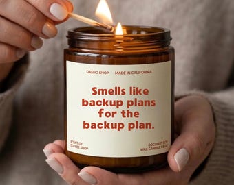 Peut inclure: Une bougie en pot de verre brun avec une étiquette crème portant le texte "Smells like backup plans for the backup plan." L'étiquette indique également "DASHO SHOP," "MADE IN CALIFORNIA," "SCENT OF COFFEE SHOP," et "COCONUT SOY WAX CANDLE 212 G."