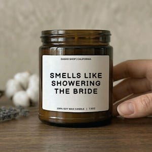 Puede incluir: Una vela en tarro de cristal marrón con una etiqueta blanca que dice "SMELLS LIKE SHOWERING THE BRIDE". La vela es de 7,5 oz y está hecha de cera de soja 100%. El texto "DASHO SHOP | CALIFORNIA" está impreso en la parte superior de la etiqueta.