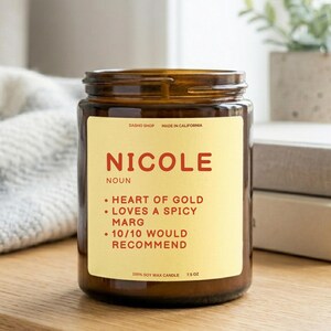 Puede incluir: Una vela de cera de soja de 213g en un frasco de vidrio ámbar con una etiqueta color crema. La etiqueta presenta el texto NICOLE NOUN, seguido de tres puntos: HEART OF GOLD, LOVES A SPICY MARG y 10/10 WOULD RECOMMEND. También indica DASHO SHOP.