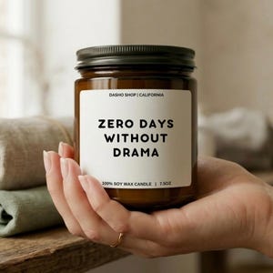 Puede incluir: Una vela en tarro de cristal marrón con tapa negra, sostenida en una mano. La etiqueta dice "ZERO DAYS WITHOUT DRAMA" en letras negras en negrita. La vela es de 213 gramos y está hecha de cera de soja 100%.