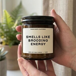 Puede incluir: Una vela en tarro de cristal marrón con tapa negra. La etiqueta dice "SMELLS LIKE BROODING ENERGY" y "100% SOY WAX CANDLE | 7.5OZ". El texto "DASHO SHOP | CALIFORNIA" está en la parte superior de la etiqueta.