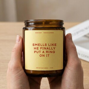 Puede incluir: Una vela de cera de soja de 7,5 oz en un frasco de vidrio ámbar oscuro con una etiqueta rectangular color crema. La etiqueta presenta el texto "DASHO SHOP MADE IN CALIFORNIA" en la parte superior, "SMELLS LIKE HE FINALLY PUT A RING ON IT" en rojo y "100% SOY WAX CANDLE 7.5 OZ" debajo.