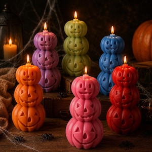 May include: A collection of six stacked pumpkin-shaped candles in orange, purple, green, blue, pink, and red. Each candle features three carved jack-o'-lantern faces with lit wicks at the top, creating a vibrant display of colorful wax figures.