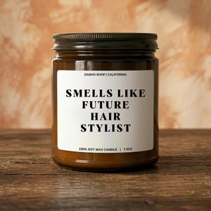 Pode incluir: Uma vela de pote de vidro castanho com tampa preta. O rótulo diz "SMELLS LIKE FUTURE HAIR STYLIST". A vela tem 213 gramas e é feita de cera de soja 100%. O fundo é de uma cor quente e neutra.