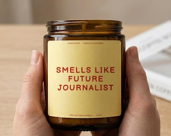 Peut inclure: Une bougie en pot de verre brun avec une étiquette jaune portant le texte rouge "SMELLS LIKE FUTURE JOURNALIST". L'étiquette affiche également "DASHO SHOP", "MADE IN CALIFORNIA", "100% SOY WAX CANDLE" et "212 G" en plus petits caractères rouges.