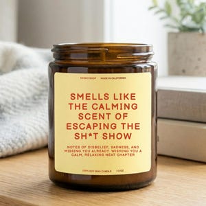 Puede incluir: Una vela en frasco de vidrio marrón con una etiqueta color crema que presenta el texto: SMELLS LIKE THE CALMING SCENT OF ESCAPING THE SH*T SHOW. La etiqueta incluye el texto: DASHO SHOP, MADE IN CALIFORNIA, 100% SOY WAX CANDLE, 212 G, Y NOTAS.
