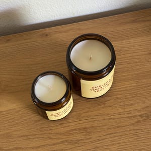 Peut inclure: Deux bougies en pot en verre ambr&eacute; avec de la cire blanche et une m&egrave;che chacune. Les bougies pr&eacute;sentent une &eacute;tiquette de couleur cr&egrave;me avec le texte "SMELLS LIKE YOUR CUSTOM TEXT HERE" imprim&eacute; en police sans-serif marron fonc&eacute; sur le devant.