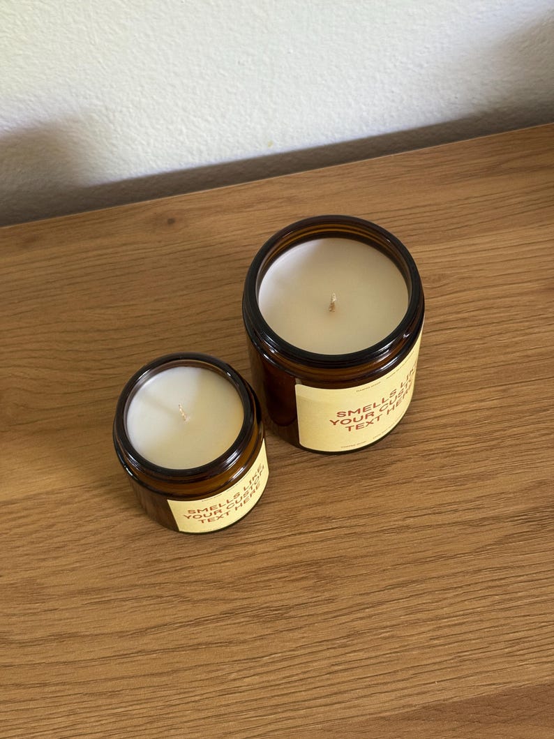 Peut inclure: Deux bougies en pot en verre ambr&eacute; avec de la cire blanche et une m&egrave;che chacune. Les bougies pr&eacute;sentent une &eacute;tiquette de couleur cr&egrave;me avec le texte "SMELLS LIKE YOUR CUSTOM TEXT HERE" imprim&eacute; en police sans-serif marron fonc&eacute; sur le devant.