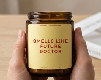 Peut inclure: Une bougie en pot de verre brun avec une étiquette jaune portant le texte rouge "SMELLS LIKE FUTURE DOCTOR". L'étiquette indique également "DASHO SHOP", "MADE IN CALIFORNIA", "100% SOY WAX CANDLE" et "212g" en petites lettres noires.