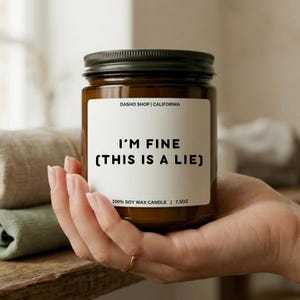 May include: A brown glass candle jar with a black lid, held in a hand. The label reads "I'M FINE (THIS IS A LIE)" and "100% SOY WAX CANDLE | 7.5OZ". The text "DASHO SHOP | CALIFORNIA" is at the top of the label.