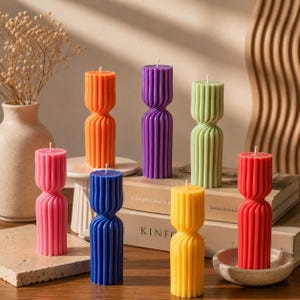 May include: A collection of seven pillar candles in vibrant colors including orange, purple, light green, pink, blue, yellow, and red. Each candle features a distinct vertical ribbed texture with a narrow, twisted waist design and a single wick.