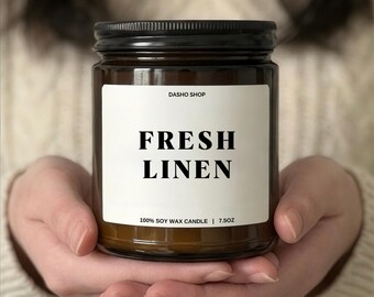 May include: A brown glass jar candle with a black lid and a white label. The label features the text DASHO SHOP, FRESH LINEN, and 100% SOY WAX CANDLE | 7.5OZ in black lettering. The candle is held in place, showcasing the clear glass and label.