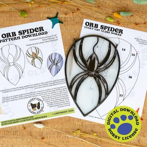 May include: A stained glass orb spider with black glass legs and body, positioned over a printed paper pattern titled Orb Spider Pattern Download. The pattern includes instructions, numbered pieces, and the Lucky Cat Glass Works logo.